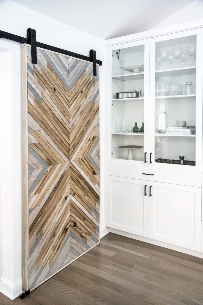 Whole-house remodel: Custom built chevron-pattern wood barn door on black track beside white glass-front built-in cabinets