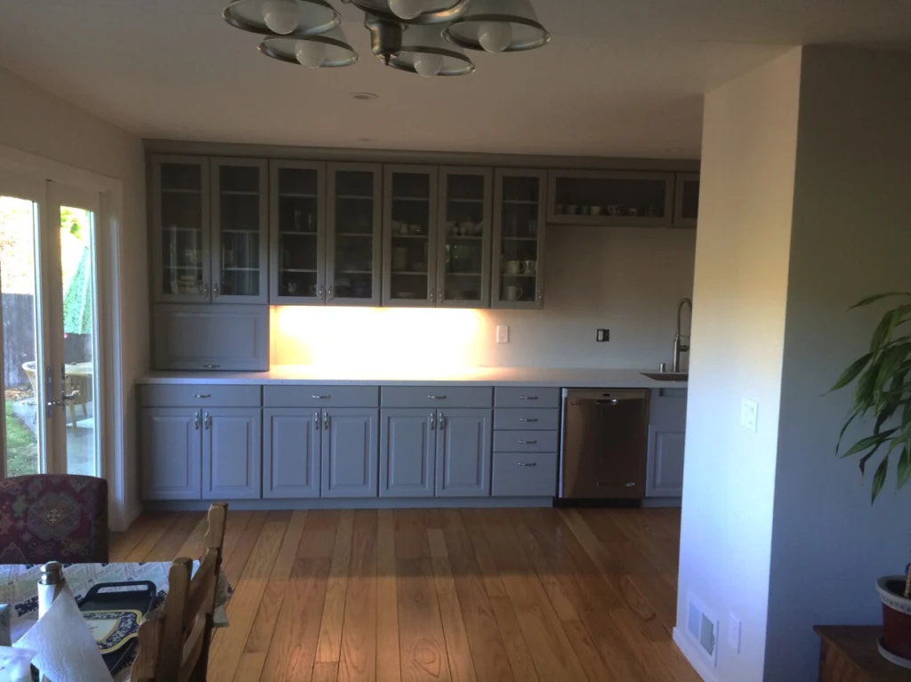 Flooring: Wall of grey cabinets with glass-front uppers, under-cabinet lighting, stainless dishwasher and oak floor