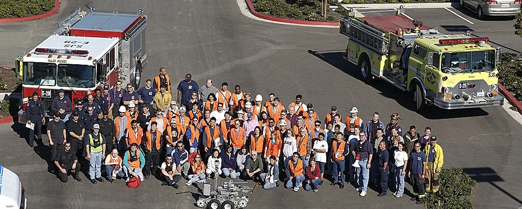Group photo of volunteers in orange vests and firefighters lined up between two fire engines; John Leimer is in the front row, right