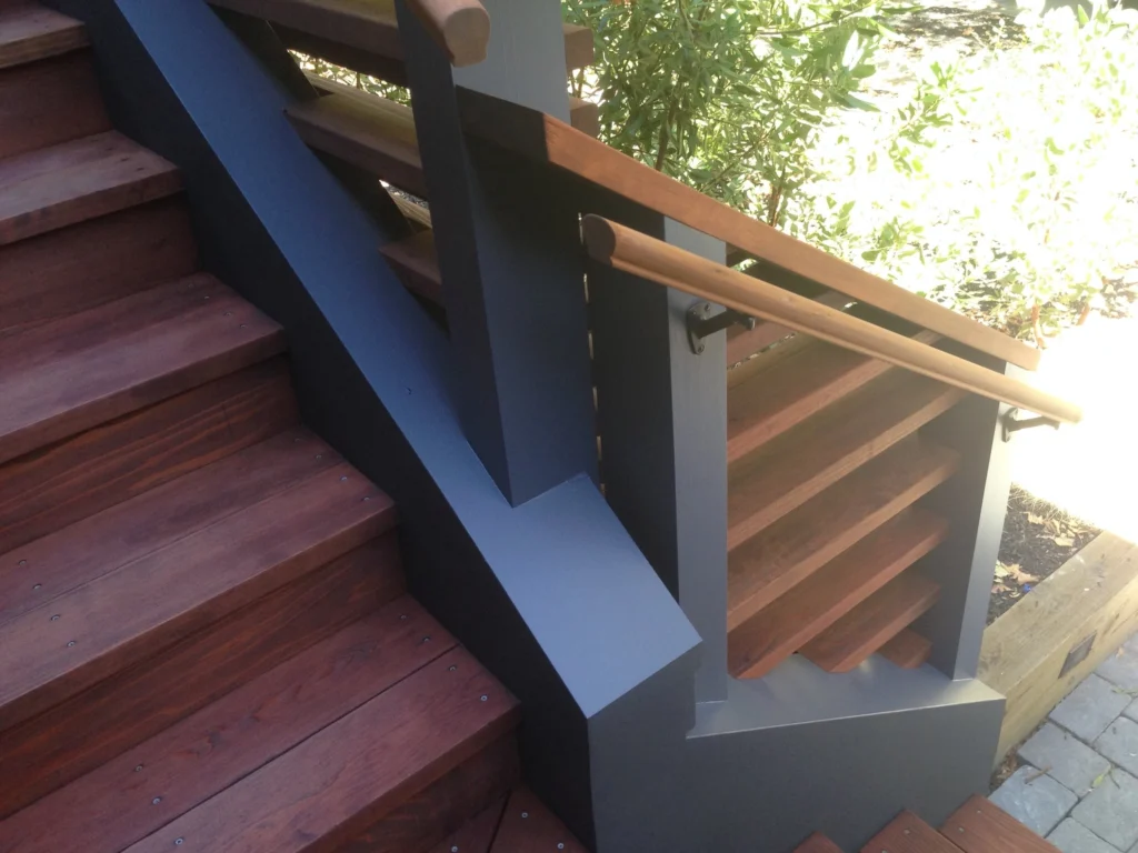 Deck: Close-up of stained wood stair treads, grey painted stringer and posts, and a round wood handrail